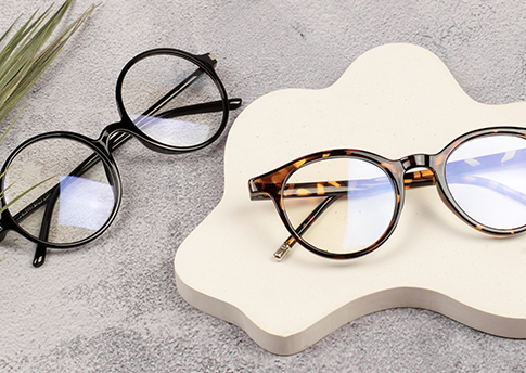 Dos gafas graduadas, una de montura redonda negra y otra de montura redonda carey, colocadas sobre una superficie gris y una base decorativa en forma orgánica.